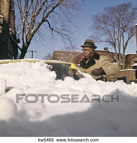 Stock Photography - 1970s AFRICAN AMERICAN MAN CLEANING SNOW ICE FROM CAR WINDSHIELD. Fotosearch