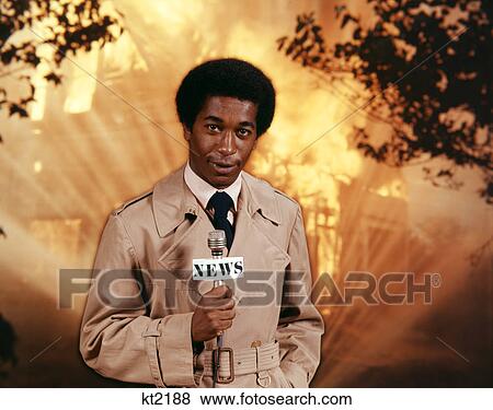 Stock Photo - 1970s AFRICAN AMERICAN TV NEWSMAN WEARING TRENCH COAT LOOKING AT CAMERA HOLD MICROPHONE REPORTING AT SCENE OF RAGING HOUSE FIRE. Fotosearch
