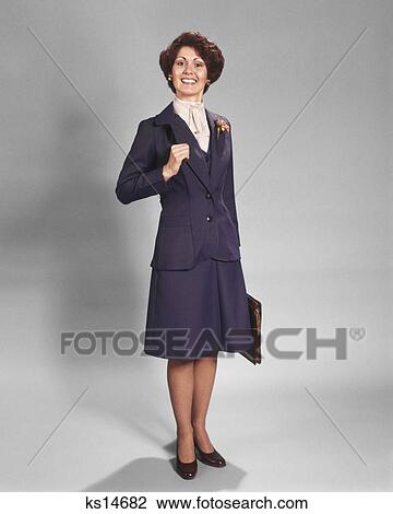 Stock Image - 1970s CONFIDENT HAPPY SMILING BUSINESSWOMAN STANDING WEARING BLUE BUSINESS SUIT HOLDING BRIEFCASE LOOKING AT CAMERA. Fotosearch