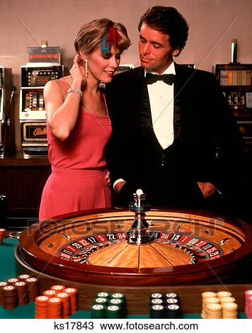 Stock Image - 1970S Couple In Formal Dress Man Tuxedo Woman Pink Dress And Diamonds Standing By Casino Roulette Wheel. Fotosearch