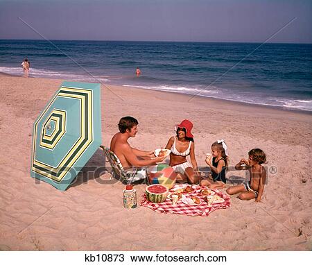 Stock Image - 1970s FAMILY EATING PICNIC AT OCEAN BEACH. Fotosearch