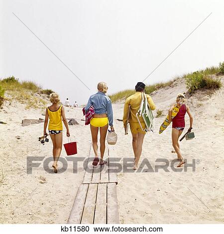 Stock Image - 1970s FAMILY MOTHER FATHER TWO BOYS WALKING ON TO BEACH CARRYING CHAIRS UMBRELLA GEAR. Fotosearch
