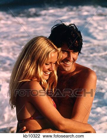 Stock Image - 1970S Happy Couple Beach Hugging Embracing Hug Embrace Laughing Blond Woman Brunette Man. Fotosearch
