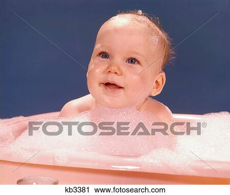 Stock Image - 1970s HAPPY SMILING BABY BATHING IN PINK PLASTIC BATHTUB SURROUNDED BY SOAP SUDS. Fotosearch