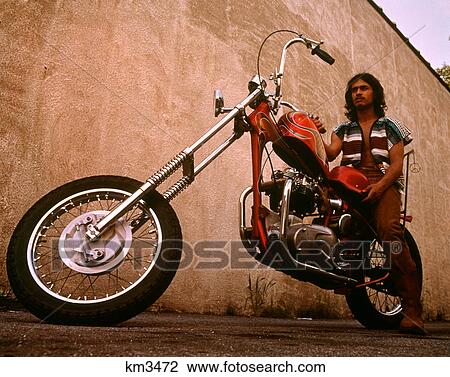 Stock Image - 1970s MAN HISPANIC HIPPIE WEARING SERAPE FABRIC VEST SITTING ON CHOPPER MOTORCYCLE. Fotosearch