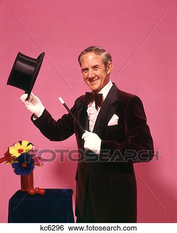 Stock Photograph - 1970s MAN MAGICIAN LOOKING AT CAMERA TIPPING TOP HAT WITH WHITE GLOVES BOW TIE MAGIC WAND FLOWERS. Fotosearch