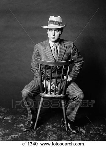 Stock Image - 1970S Man Seated Backwards On Chair Looking At Camera Holding Gun Wearing Slick Stylish Pinstripe Suit And Hat. Fotosearch