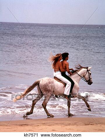 Stock Image - 1970s MAN WOMAN COUPLE RIDING TOGETHER ON SINGLE WHITE GRAY HORSE ON BEACH. Fotosearch