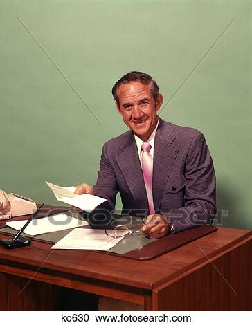 1970s MIDDLE-AGED MAN SITTING AT DESK HOLDING CHECK SMILING LOOKING AT CAMERA View Large Photo Image Stock Image - 1970s MIDDLE-AGED MAN SITTING AT DESK HOLDING CHECK SMILING LOOKING AT CAMERA. Fotosearch