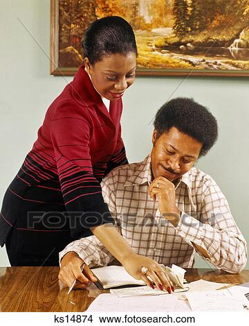 Picture - 1970s SMILING AFRICAN AMERICAN COUPLE AT TABLE WRITING CHECKS PAYING BILLS. Fotosearch
