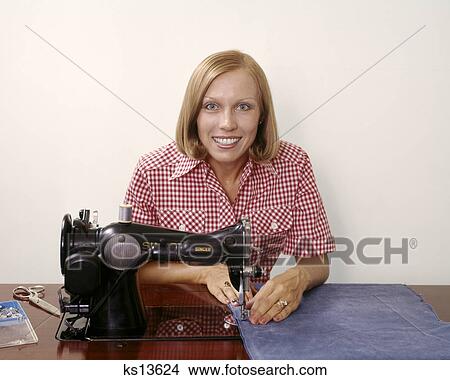 Picture - 1970s SMILING BLOND WOMAN HOUSEWIFE LOOKING AT CAMERA OPERATING ANTIQUE BLACK SINGER SEWING MACHINE. Fotosearch