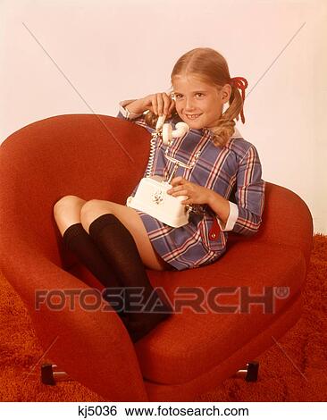 Stock Photograph - 1970S Smiling Girl Knee Socks Plaid Dress Sitting Red Chair Talking Phone Telephone. Fotosearch