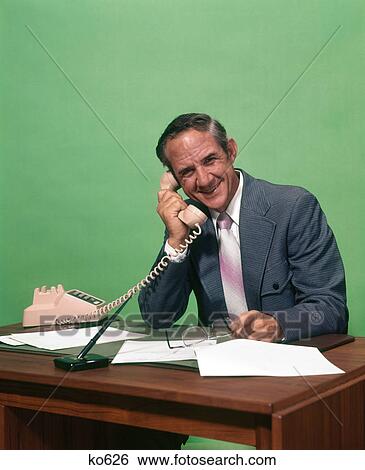 1970s SMILING MATURE MAN WEARING SUIT HOLDING GLASSES SITTING AT DESK SPEAKING ON TELEPHONE LOOKING AT CAMERA View Large Photo Image Stock Photograph - 1970s SMILING MATURE MAN WEARING SUIT HOLDING GLASSES SITTING AT DESK SPEAKING ON TELEPHONE LOOKING AT CAMERA. Fotosearch