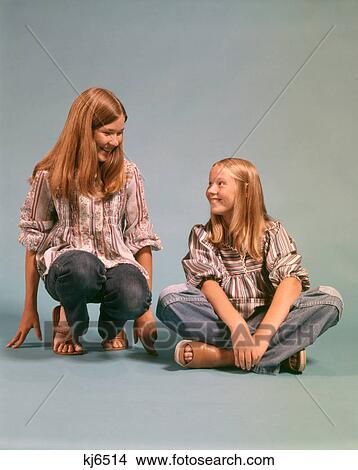 1970s TWO TEEN GIRLS SISTERS WEARING BLUE JEANS DENIM PEASANT SHIRTS SITTING AND KNEELING SMILING LOOKING AT EACH OTHER View Large Photo Image Picture - 1970s TWO TEEN GIRLS SISTERS WEARING BLUE JEANS DENIM PEASANT SHIRTS SITTING AND KNEELING SMILING LOOKING AT EACH OTHER. Fotosearch