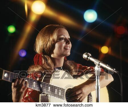 Stock Photo - 1970s WOMAN FOLK SINGER GUITAR SINGING INTO MICROPHONE COLORFUL SPOTLIGHTS. Fotosearch
