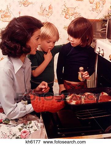 Stock Photo - 1970S Woman Mother 2 Children Boy Girl Kitchen Oven Door Open Checking Steaks Meat Adding Spice Cooking. Fotosearch