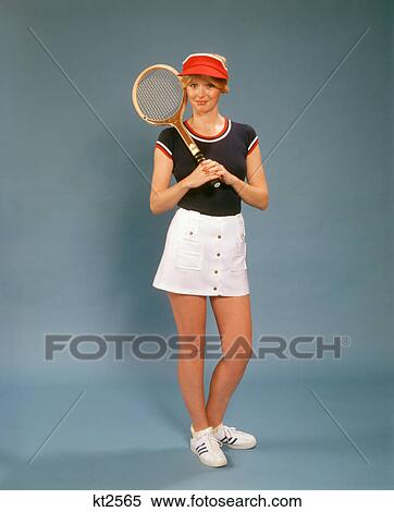 Stock Photography - 1970s WOMAN ON BLUE SEAMLESS HOLDING TENNIS RACKET WEARING SHORT WHITE SKIRT RED VISOR BLUE TEE LOOKING AT CAMERA. Fotosearch