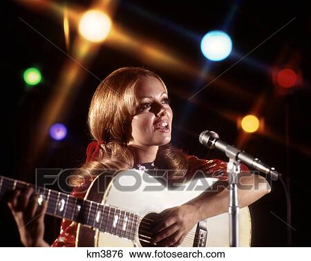 Stock Photograph - 1970s WOMAN PLAYING GUITAR SINGING INTO MICROPHONE COLORED SPOTLIGHTS. Fotosearch