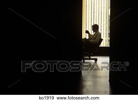 Stock Photo - 1980s ANONYMOUS SILHOUETTED YOUNG BUSINESS MAN DRINKING COFFEE SEATED AT DESK SEEN THROUGH OFFICE DOOR. Fotosearch