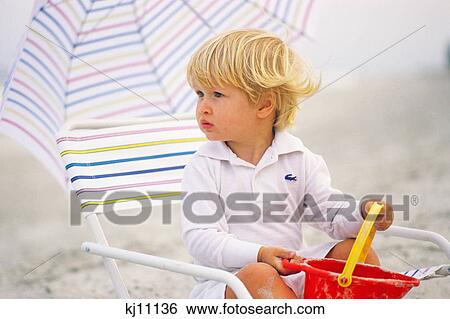 1980s BLONDE BOY TODDLER SITTING IN BEACH CHAIR HOLDING SAND PAIL View Large Photo Image Stock Photograph - 1980s BLONDE BOY TODDLER SITTING IN BEACH CHAIR HOLDING SAND PAIL. Fotosearch