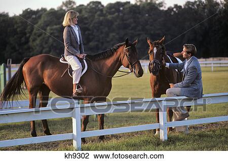 Stock Image - 1980s COUPLE WITH HORSES WOMAN SITTING ON HORSE MAN ON FENCE TALKING. Fotosearch