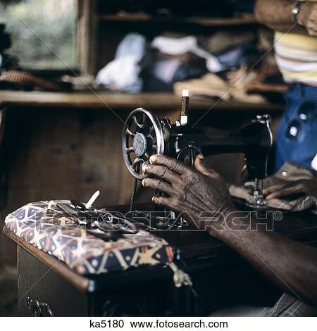 Stock Image - 1980s HAND OF OLD AFRICAN AMERICAN WOMAN SEWING ON ANTIQUE CAST IRON SEWING MACHINE. Fotosearch