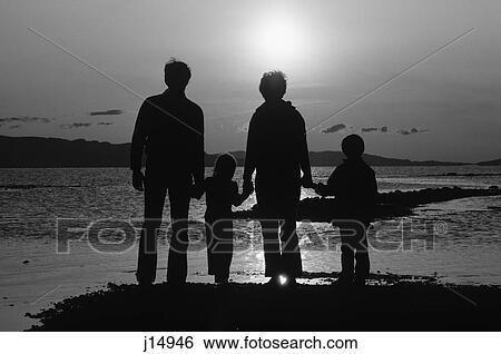 Stock Photograph - 1980s REAR VIEW SILHOUETTE OF ANONYMOUS FAMILY OF FOUR STANDING ON BEACH HOLDING HANDS AT SUNSET. Fotosearch