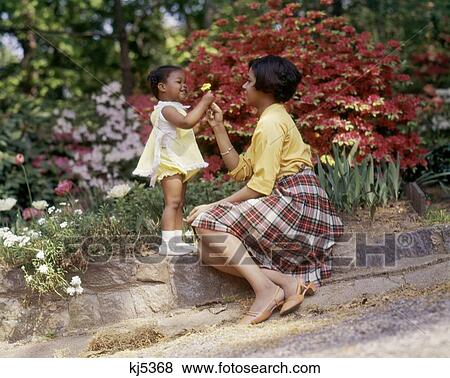 Stock Photo - 1980S Smiling African American Woman And Little Girl Outdoors Near Spring Flowers Daughter Giving Yellow Flower To Mother. Fotosearch