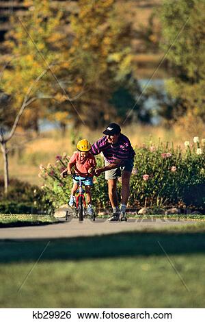 Stock Photograph - 1990s FATHER TEACHING CHILD WEARING YELLOW HELMET TO RIDE BICYCLE WITH TRAINING WHEELS. Fotosearch