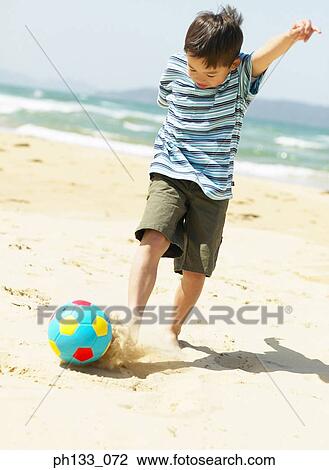 Stock Image - Young boy playing football on beach. Fotosearch