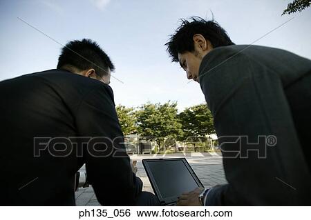 Stock Photograph - Rear view of two young businessmen working outdoors. Fotosearch