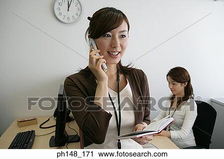 Close-up of a businesswoman talking on a mobile phone with her colleague using a computer in the background View Large Photo Image Stock Image - Close-up of a businesswoman talking on a mobile phone with her colleague using a computer in the background. Fotosearch