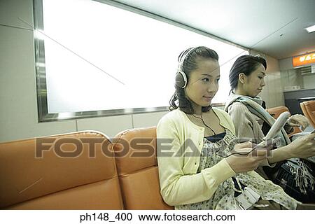 Close-up of a young woman listening to music on headphones with her friend reading a newspaper View Large Photo Image Stock Image - Close-up of a young woman listening to music on headphones with her friend reading a newspaper. Fotosearch