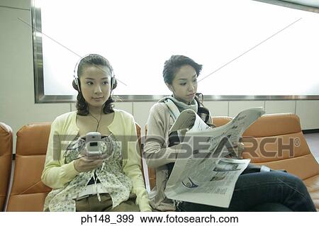 Close-up of a young woman listening to music on headphones with her friend reading a newspaper View Large Photo Image Stock Photo - Close-up of a young woman listening to music on headphones with her friend reading a newspaper. Fotosearch