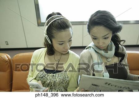 Close-up of a young woman listening to music on headphones with her friend reading a newspaper View Large Photo Image Stock Photograph - Close-up of a young woman listening to music on headphones with her friend reading a newspaper. Fotosearch
