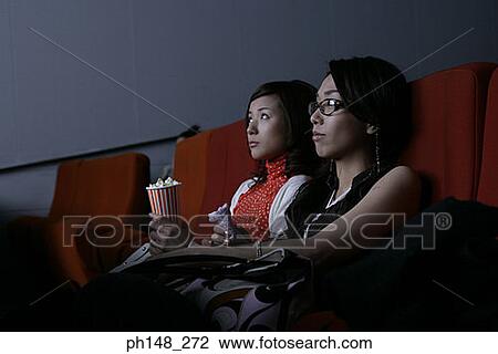 Stock Image - Close-up of two young women watching a movie in a movie theater. Fotosearch