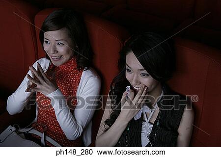 Stock Image - High angle view of two young women watching a movie in a movie theater. Fotosearch