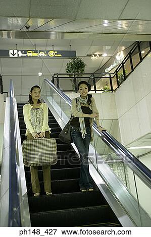 Stock Photo - Low angle view of two young women standing on an escalator and smiling. Fotosearch