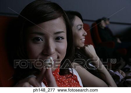 Stock Photo - Portrait of two young women eating popcorn and watching a movie in a movie theater. Fotosearch