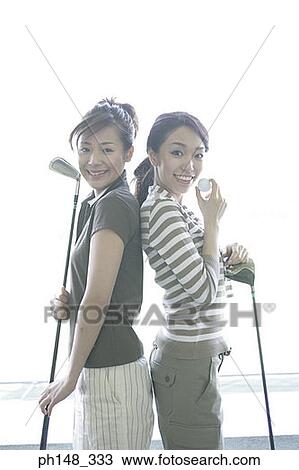 Stock Image - Portrait of two young women standing back to back and holding golf clubs. Fotosearch