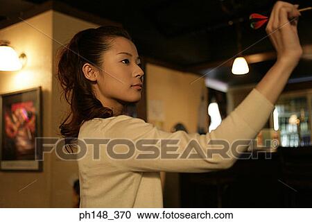 Side profile of a young woman holding a dart and aiming at a dart board in a nightclub View Large Photo Image Stock Image - Side profile of a young woman holding a dart and aiming at a dart board in a nightclub. Fotosearch