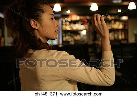 Side profile of a young woman holding a dart and aiming at a dart board in a nightclub View Large Photo Image Stock Image - Side profile of a young woman holding a dart and aiming at a dart board in a nightclub. Fotosearch