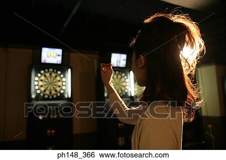 Stock Photograph - Side profile of a young woman holding a dart and aiming at a dart board in a nightclub. Fotosearch
