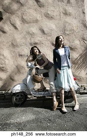 Stock Photograph - Side profile of a young woman sitting on a scooter with her friend standing beside her. Fotosearch