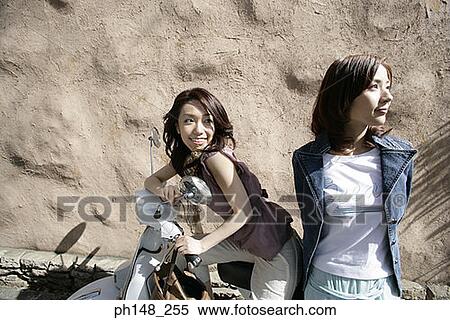 Stock Photography - Side profile of a young woman sitting on a scooter with her friend standing beside her. Fotosearch