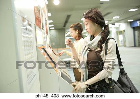 Stock Photo - Side profile of two young women operating a touch screen at a subway station. Fotosearch