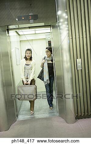 Stock Photo - Two young women walking out of an elevator with luggage. Fotosearch