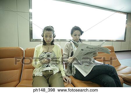 Stock Photo - Young woman listening to music on headphones with her friend reading a newspaper. Fotosearch
