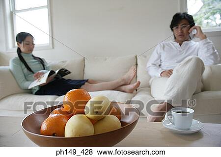 Stock Photo - Close-up of a fruit bowl on the table with a young couple sitting on a couch in the background. Fotosearch