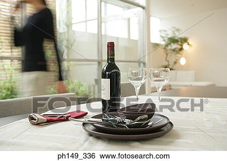Stock Photograph - Close-up of crockery and a wine bottle in a table with a woman in the background. Fotosearch